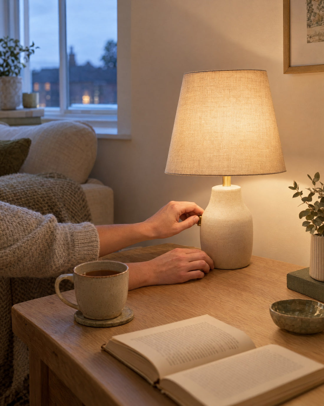 Hands switching on a linen-shade table lamp beside a cup of tea and an open book