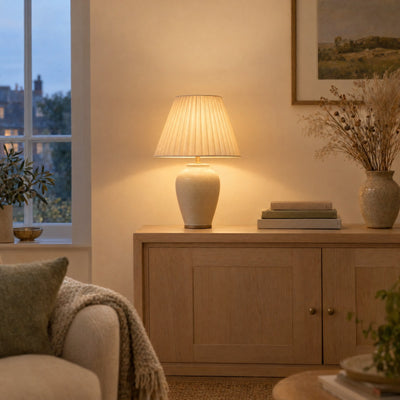 Ceramic table lamp with a pleated shade glowing on an oak sideboard