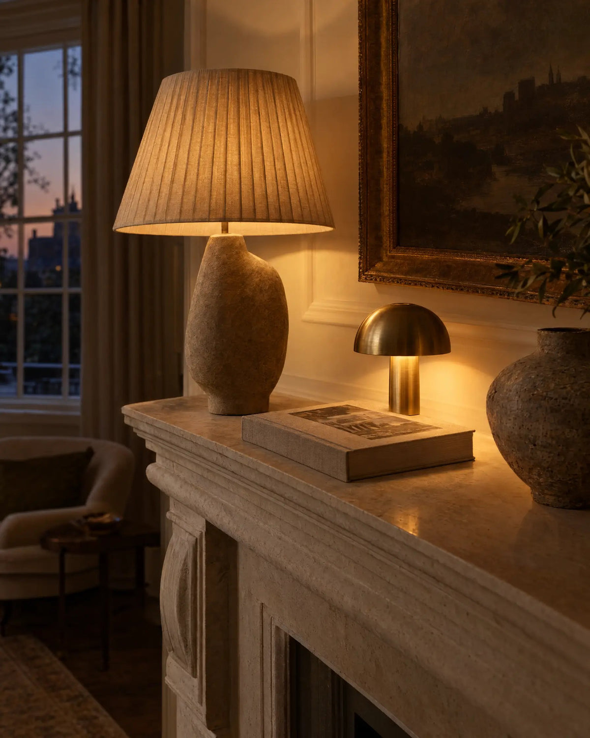 Pleated linen table lamp and cordless brass accent lamp glowing warmly on a limestone mantelpiece at dusk